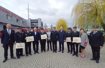 Jedenastu mężczyzn i kobieta. Ubrani na galowo/mundurach strażackich. W tle budynek straży pożarnej 